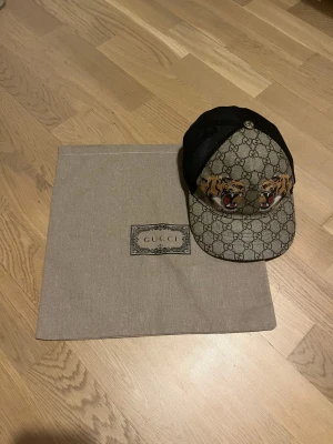 Gucci keps med tigertryck och mesh - Snygg Gucci keps med klassiskt GG-mönster i beige och brunt, prydd med två broderade tigrar framtill. Baksidan är i svart mesh för extra ventilation och har justerbar rem med röd-gröna detaljer. Den är i storlek S dvs 57cm och det finns inga defekter alls endast att formen måste fixas litegrann.