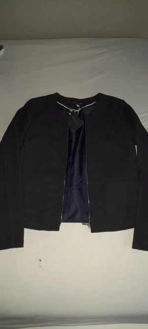 Svart kavajjacka från Premium Selection - Svart kavajjacka med rund hals och raka linjer från Premium Selection. Jackan har två stora fickor fram, hel dragkedja och är helfodrad med mörkblått tyg. Materialet är mjukt och har en lätt struktur, perfekt för en clean och stilren look.