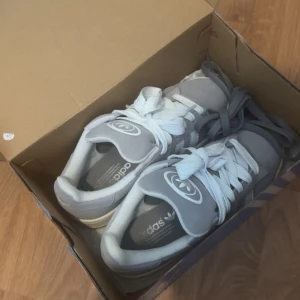 Adidas Campus grå sneakers mocka - Säljer ett par Adidas Campus sneakers i grå mocka med vita detaljer och klassiska tre ränder på sidan. Skorna har rund tå, platt sula och snörning med både vita och grå skosnören. Adidas-logga på plösen och sidan. Perfekt för dig som gillar stilrena och tidlösa sneakers.