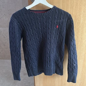 Mörkblå kabelstickad tröja Polo Ralph Lauren - Kabelstickad mörkblå tröja från Polo Ralph Lauren med klassisk rund hals och röd broderad logga på bröstet. Tröjan har ribbade muddar vid ärmslut och nederkant, samt långärmad passform. Perfekt för dig som gillar stilrena och tidlösa plagg. Lite urtvättat 