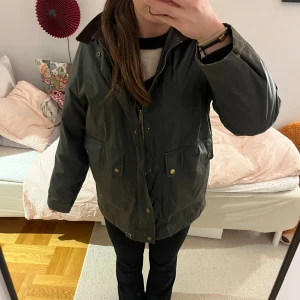 Mörkgrön parkajacka med brunt krage - Säljer en klassisk mörkgrön parkajacka från Zara med brun krage, stora fickor med tryckknappar och dragkedja framtill. Jackan har en loose passform och är gjord i polyester med vaxad yta som ger en cool look. Perfekt för höst och vinter. Jackan saknar en knapp till fickorna på framsidan och är lite nopprig innuti.