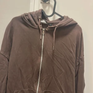 Brun hoodie med dragkedja - Mjuk brun hoodie med huva och snörning, tillverkad i sammet. Tröjan har en hel dragkedja framtill och praktiska fickor. Perfekt för chill dagar och enkel att matcha med jeans eller mjukisbyxor.