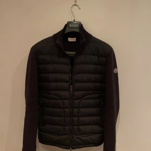 Moncler cardigan  - Svart dunjacka från Moncler med quiltad front och stickade ärmar. Jackan har dragkedja framtill, två sidofickor med dragkedja och Moncler-logga på vänster ärm. Snygg, stilren och perfekt för kyliga dagar. Priset är inte hugget i sten!