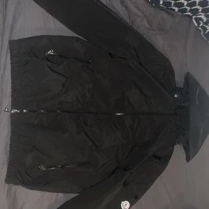 Svart dunjacka från Moncler - Svart dunjacka från Moncler med huva och två dragkedjeförsedda fickor framtill. Jackan har en clean look med Moncler-logga på ärmen och DIST-certifierad dunfyllning. Perfekt för dig som vill ha en stilren jacka med premiumkänsla.