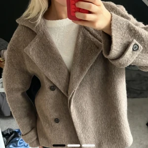 Beige ullkappa från Arket, strl 38 - Säljer en snygg beige kappa från Arket i storlek 38. Jackan är tillverkad i ullblandning, har bred krage, dubbelknäppning framtill och knappar vid ärmsluten. Perfekt för höst och vinter, med en loose fit och stilren look.