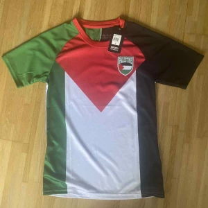 Palestina fotbollströja med emblem - Snygg fotbollströja inspirerad av Palestinas flagga med emblem på bröstet. Tröjan har korta ärmar och är tillverkad i ett lätt, syntetiskt material som andas. Färgerna är röd, grön, vit och svart i ett grafiskt mönster. Perfekt för match eller träning.