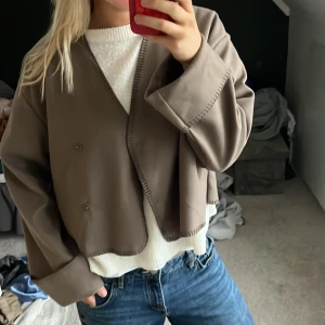 Brun croppad jacka från Gina Tricot - Säljer en brun croppad jacka från Gina Tricot med oversized passform och snygga synliga sömmar. Jackan har omlottknäppning med knappar och långa ärmar. Perfekt för dig som gillar en stilren och modern look.