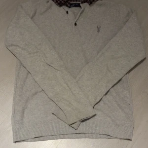 Grå pikétröja med rutig krage från Next - Snygg grå pikétröja från Next med långärmad design och rutig krage i svart, vit och röd. Tröjan har knappar vid halsen och ett diskret broderat emblem på bröstet. Perfekt för dig som gillar klassisk stil med en twist. Priset kan diskuteras vid snabb affär!