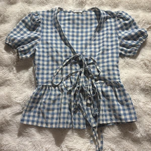 Blårutig blus med snörning från smileville - Supersöt blus i blåvitt rutigt från smileville med puffiga korta ärmar och omlott-design. Framtill finns flera snörningar som ger en unik look. Blusen har volangkant nedtill och v-ringad hals. Perfekt för dig som gillar romantiska och trendiga detaljer.