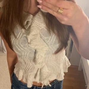 Vit omlott blus med volanger  - Säljer en vit blus med snygga volanger framtill och omlott-design. Blusen har ett lätt krinklat tyg och är ärmlös, vilket ger en cool och feminin vibe. Perfekt att styla med jeans eller kjol för en trendig look.