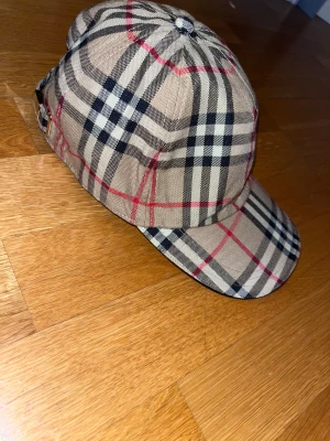 Burberry rutig keps i bomull - Snygg Burberry keps med klassiskt rutigt mönster i beige, svart och rött. Justerbar rem baktill med metallspänne. Tillverkad i bomull och har den ikoniska Burberry-looken som lyfter din stil till nästa nivå.