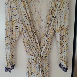 Blommig morgonrock/negligé Calvin Klein XS/S - Snygg morgonrock/ negligé från Calvin Klein med blommigt mönster i ljusa toner, inslag av gult, lila och grönt. Långärmad med spetsdetaljer vid ärmslut och knyt i midjan. Tillverkad i mjuk modal som känns skön mot huden.