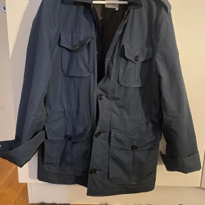 Marinblå parkajacka från J.Lindeberg - Parkajacka från J.Lindeberg i marinblå färg med fyra stora fickor framtill och snygga knappar. Jackan har huva och en metallbadge på ärmen med märkets logga. Perfekt för dig som gillar stilren och funktionell design.