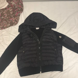 Svart Moncler pufferjacka med huva - Svart Cardigan från Moncler med stickade ärmar och quiltad kropp. Jackan har en stor huva, dragkedja framtill och en Moncler-logga på ärmen. Materialmix av polyester och stickad textil ger en schysst kontrast. Perfekt för kyliga dagar.