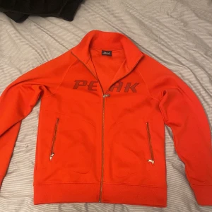 Röd ziptröja Peak Performance  - Ny pris, 2200 kr. Snygg röd ziptröja från Peak Performance med broderad PEAK-logga på bröstet. Den är storlek L men passar perfekt till de som har storlek M. Tröjan har hög krage, två fickor med dragkedja och hel dragkedja framtill. Perfekt för lager-på-lager och chill dagar. Materialet är mjukt och känns som bomull. Det är passande till lite vintage stil och den är unik!