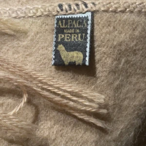 Beige alpackahalsduk  - Snygg beige halsduk i mjuk alpackaull från Peru. Den är enfärgad och har långa, tvinnade fransar i ändarna. Perfekt att svepa runt halsen för extra värme och stil. Klassisk rektangulär form och diskret söm längs kanterna.