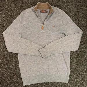 Ljusblå merinoulltröja från Morris - Snygg ljus blå stickad tröja från Morris i 100% merinoull. Tröjan har en half zip med läderdetalj, ribbad krage i beige och långärmad passform. Diskret Morris-logga broderad på bröstet. Perfekt för dig som gillar stilrena och bekväma plagg.