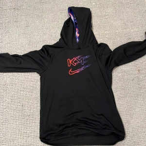 Nike svart hoodie med flames - Svart hoodie från Nike med Dri-Fit material, perfekt för träning. Framsidan har KM-logga och Nike Swoosh i rött och lila med flames. Insidan av huvan har ett blått och lila flame-mönster. Stor ficka framtill och långa ärmar. Snygg och sportig look! Från kylian mbappes egna kollektion, köpt för ca 700-800 kr.