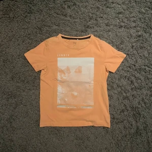 Persikofärgad t-shirt från Name It 9-10år - Snygg persikofärgad t-shirt från Name It med tryck framtill som visar ett strandmotiv och texten 'SUMMER'. T-shirten har rund hals och korta ärmar, tillverkad i mjuk ekologisk bomull. Perfekt för dig som gillar chill och sommarkänsla. Passar 9-10 år. Storlek 134 - 140