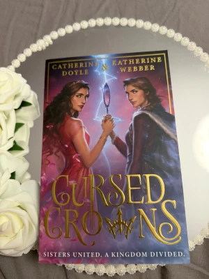 Cursed Crowns - En magisk och spännande bok med två systrar i centrum, där mystik och kungliga intriger möts. Perfekt för dig som gillar fantasy, starka karaktärer och episka äventyr. Omslaget lockar med gnistrande detaljer och en dramatisk stämning.