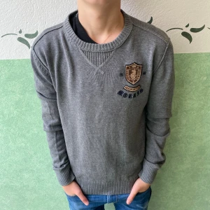 Grå stickad tröja från Morris - Snygg grå stickad tröja från Morris med broderad logga och emblem på bröstet. Tröjan har rund hals, ribbade muddar och lång ärm. Perfekt för dig som gillar klassisk stil med en sportig touch. Passar dig som vill ha en normal passform.