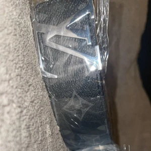  svart bälte med monogram - Svart bälte från  med klassiskt monogrammönster och stor spänne i silver. Bältet är gjort i präglat skinn och har en lyxig känsla med tydliga detaljer och ikonisk design.