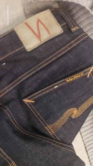 Mörkblå Nudie Jeans 32/34 - Säljer ett par mörkblå jeans från Nudie Jeans i klassisk rak modell. Jeansen har orange kontrastsömmar, fem fickor och Nudie-logga på bakfickan. Tillverkade i slitstarkt bomullsjeansmaterial. Perfekt för dig som gillar stilrena och tidlösa jeans.