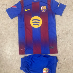 FC Barcelona Raphinha matchtröja - Säljer en FC Barcelona matchtröja med Raphinha och nummer 11 på ryggen. Tröjan är kortärmad, har klassiska blå och vinröda ränder, gult Nike-märke och klubbmärke på bröstet. Materialet är Dri-FIT polyester, perfekt för fotboll. Shorts med klubbmärke ingår.