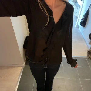 Svart volangblus från Anna Field - Säljer en svart blus från Anna Field med långa ärmar och snygga volangdetaljer längs knäppningen och vid ärmsluten. Blusen är gjord i ett lätt och transparent material som ger en luftig känsla och har en klassisk krage.