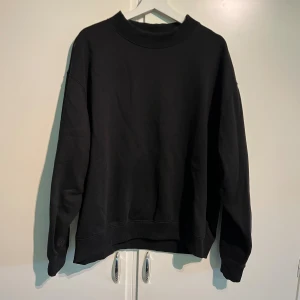 Svart sweatshirt från Weekday - En svart sweatshirt från Weekday i storlek L. Tröjan har en klassisk rund halsringning med ribbade muddar vid hals, ärmslut och nederkant. Oversized passform som ger en avslappnad vibe. Perfekt att slänga på sig när du vill vara både bekväm och stilren. Köpt för ca 1 år sedan och använd fåtal gånger, nypris runt 600-800kr