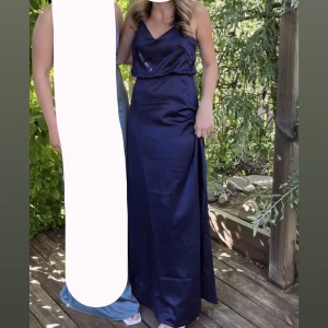 Marinblå balklänning i satin - Säljer min balklänning. Snygg marinblå balklänning i glansigt satinmaterial med v-ringad hals och smala axelband. Klänningen har en elegant och enkel siluett som ger en stilren look. Perfekt för dig som vill sticka ut med en klassisk och tidlös stil. Uppsydd, men väldigt lätt att sprätta upp. Använd 1 gång. Är 1,63, hade 6cm klackar och de nuddar backen