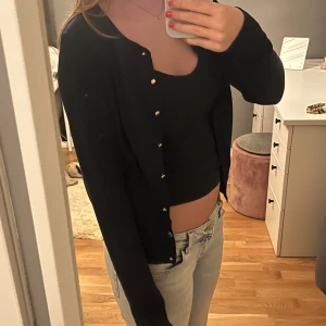 Svart kofta från Holly & Whyte - Klassisk svart kofta från Holly & Whyte by Lindex med rund halsringning och snygga guldfärgade knappar framtill. Långärmad och i mjuk bomullskvalitet, perfekt att slänga över ett linne eller t-shirt. Enkel och stilren design som funkar till allt.