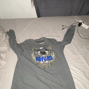 Grå Kenzo tröja med tigertryck - Säljer en grå sweatshirt från Kenzo med ikoniskt broderat tigerhuvud och blått Kenzo-tryck på bröstet. Tröjan har rund hals, långa ärmar och är gjord i mjuk bomull. Perfekt för dig som vill sticka ut med en klassisk streetstyle-look. Jag har S och den passar perfekt för mig. Lappen gick från tröjan men skickar med den i posten