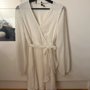 Vit omlottklänning från Vero Moda - Superfin vit omlottklänning från Vero Moda med knytband i midjan och diskret prickig struktur. Klänningen har långa ärmar med resår vid handlederna och en lätt volangkant nertill. Perfekt för sommarens alla tillfällen!
