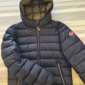 Mörkblå Colmar pufferjacka med huva - Säljer en mörkblå pufferjacka från Colmar med huva och dragkedja framtill. Jackan har två snedställda fickor med dragkedja och ett broderat Colmar-märke på ärmen. Insidan är olivgrön och jackan är vadderad för extra värme. Jätte fin jacka