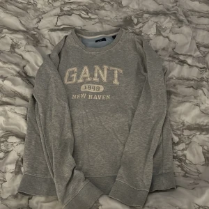 Grå sweatshirt från GANT med tryck - Snygg grå sweatshirt från GANT med stort tryck på bröstet där det står 'GANT 1949 NEW HAVEN'. Tröjan har rund hals, långa ärmar och ribbade muddar vid ärmslut och nederkant. Perfekt för dig som gillar klassisk och sportig stil.