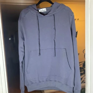 Blå hoodie från Stone Island - Säljer en stilren blå hoodie från Stone Island med huva och dragsnören. Tröjan har en klassisk känguruficka framtill och ribbade muddar vid ärmslut och nederkant. Perfekt för dig som gillar en clean och avslappnad look.