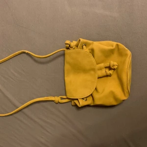 Gul mocka axelväska med snörning - Snygg gul axelväska i mjuk mocka med lång rem och praktisk snörning upptill. Väskan har ett lock som stängs framtill och en enkel, stilren design som passar perfekt till din vardagsstil.