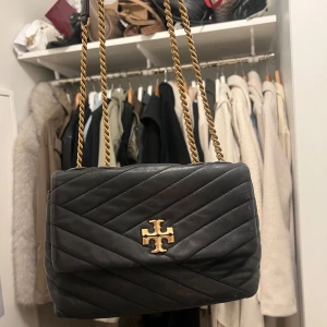 Svart quiltad axelväska Tory Burch - Säljer en svart quiltad axelväska från Tory Burch med guldfärgad kedjerem och ikonisk logga framtill. Väskan är i mjukt skinn och har ett stilrent, tidlöst uttryck. Perfekt storlek för det viktigaste och passar till många olika looks. Köpt för 7000kr säljer för 4000kr pris kan diskuteras