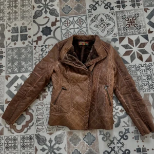 Brun quiltad skinnjacka med päls - Snygg brun skinnjacka med quiltade detaljer och mjuktpälsfoder. Jackan har dragkedja framtill och två zip-fickor. Perfekt för dig som gillar en cool och varm look med lite biker-vibe. Jackan är storlek S herr men passar kvinnor också om man vill det. Kan skicka mått om de behövs!