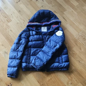 Mörkblå Moncler dunjacka med huva - Snygg mörkblå dunjacka från Moncler med huva och logotyp på ärmen. Jackan har dragkedja framtill, quiltad design och ribbade muddar. Fodrad med dun och har en sportig look med Moncler-tryck på huvan. Perfekt för kalla dagar. En del gick sönder på inner armen men har sytts så det syns ej bild nr4. 