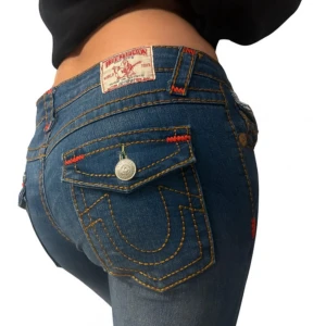 True Religion Joey jeans blå, stl 30 - Säljer ett par blå True Religion Joey jeans med ikoniska kontrastsömmar i gult och rött, stora bakfickor med lock och metallknappar. Klassisk låg midja och bootcut-modell. Tillverkade i 100% bomull, made in USA. Perfekta för dig som gillar streetwear och detaljer.