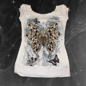 Vit topp med fjärilstryck och puffärm - Vit topp med puffärmar och ett detaljerat fjärilstryck framtill i svart, guld och beige toner. Toppen har rund halsringning och är tillverkad i mjukt bomullsmaterial. Snygga detaljer med blommor och mindre fjärilar i bakgrunden.