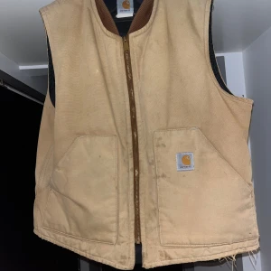 Carhartt väst - Säljer en beige väst från carhartt. Västen har en quiltad insida som gör den varm och bekväm att bära under kallare dagar och fungerar utmärkt som ytterplagg under höst och vår eller som mellanlager under vintern. Den har två stora framfickor, dragkedja framtill och den välkända Carhartt-loggan på fickan. Västen är välanvänd men i gott skick med vissa fläckar och lite slitage längst ner, vilket ger den en robust och autentisk look. Storlek saknas på etiketten men motsvarar M/L.