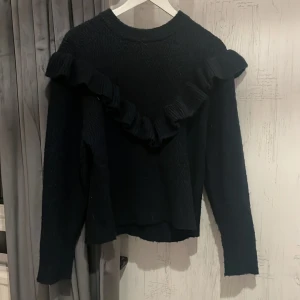 Svart stickad tröja med volang Gina Tricot - Svart stickad tröja från Gina Tricot i storlek 158/164. Tröjan har en snygg volangdetalj som går diagonalt över bröstet och axlarna. Långärmad och med rund hals. Använd fåtal gånger