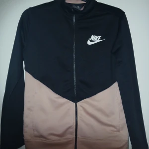 Svart och rosa Nike funktionströja - Nike funktionströja med svart överdel och rosa nedre del, klassisk logga på bröstet och hel dragkedja framtill. Tröjan har lång ärm och sportig look, perfekt för chill eller träning. Snygg färgblockad design som sticker ut. Barnstorlek 134-147 (150).