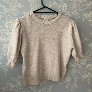 Beige stickad tröja från Gina Tricot - Superfin beige stickad tröja från Gina Tricot med puffiga korta ärmar och ribbade muddar. Tröjan har en rund halsringning och är perfekt för dig som gillar en enkel men trendig look. Passar till både jeans och kjol.