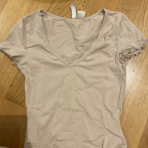 Beige v-ringad topp med kort ärm - Säljer en enkel och stilren beige topp med v-ringad hals och korta ärmar. Toppen är gjord i mjuk bomull och har en klassisk passform som är lätt att matcha med olika outfits. Perfekt för dig som gillar neutrala färger och clean look.