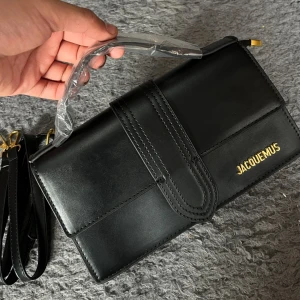 Svart handväska från Jacquemus - Snygg svart handväska från Jacquemus med stilren design och guldfärgad logga. Väskan är rektangulär, har ett avtagbart axelband och är tillverkad i slätt skinn. Perfekt för dig som gillar minimalistisk och trendig look.