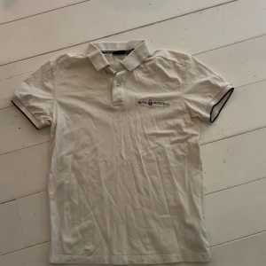 Vit pikétröja från Sail Racing - Fräsch vit pikétröja från Sail Racing med klassisk krage och två knappar framtill. Tröjan har korta ärmar med svart kant och diskret logga på bröstet. Perfekt för dig som gillar stilren och sportig look. Använd 2 gånger 